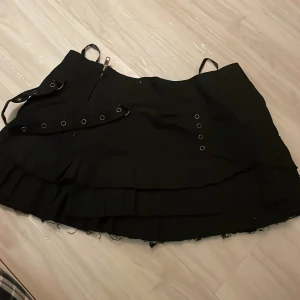 Svart kjol från Spin Doctor - Snygg svart kjol från Spin Doctor med coola detaljer som snörning och nitar. Kjolen har en volangkant och justerbara band. Perfekt för en edgy look!