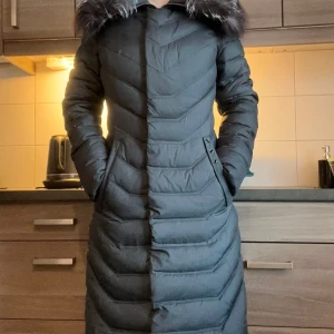 Svart dunjacka med pälskrage - Snygg svart dunjacka med pälskrage och knytband i midjan. Jackan har långa ärmar och en quiltad design som ger extra värme. Perfekt för kyliga dagar.