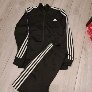 Svart träningsset från Adidas - Säljer ett svart träningsset från Adidas med de klassiska tre ränderna i vitt längs ärmar och ben. Setet består av en jacka med dragkedja och matchande byxor. Perfekt för träning eller en avslappnad dag.