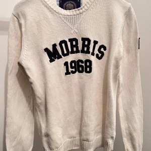 Vit stickad tröja från Morris - Säljer en stilren vit stickad tröja från Morris med texten 'Morris 1968' i svart på framsidan. Tröjan har långa ärmar och en ribbad kant vid halsen samt ärmslut och nederkant. Perfekt för en klassisk look.