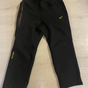 Svarta Nike Nocta byxor  - Säljer ett par svarta mjukisbyxor från Nike Nocta med gul logga och text. Byxorna har en bekväm passform med dragsko i midjan och sidofickor. ”Uncuffed hem” vid fotänden. (Dvs att den sydda delen vid fotänden är uppsprättad). Storlek L men passar som S-M