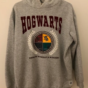 Grå Hogwarts hoodie - Säljer en grå hoodie med Hogwarts-tryck i vinrött och emblem för de fyra elevhemmen. Perfekt för alla Harry Potter-fans! Mjuk och bekväm. 