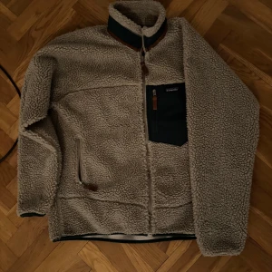 Beige fleecejacka från Patagonia - Mysig beige fleecejacka från Patagonia med dragkedja och en praktisk bröstficka. Perfekt för kyliga dagar med sin varma och mjuka design. Jackan har en hög krage och långa ärmar.