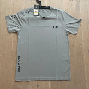 Grå t-shirt från Under Armour - Snygg grå t-shirt från Under Armour med svart logga på bröstet och text längs sidan.  Helt nya med tag och aldrig använd .