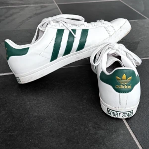 Adidas Court Star sneakers - Snygga vita Adidas Court Star sneakers med gröna detaljer och klassiska tre ränder på sidan. Skorna har snörning och guldlogga på hälen. Kika och se om du ser något mer i min profil som du gillar så skickar jag med det till dig också!😊 Fler kläder kommer! 