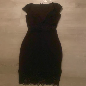 Svart klänning med spetsdetaljer från Nelly - Snygg svart bodycon-klänning från Nelly med spetsdetaljer vid fållen. Den har korta ärmar och en elegant passform som framhäver figuren. Perfekt för en kväll ute eller en speciell tillställning.