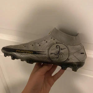 Grå Nike fotbollsskor Phantom GT - Snygga grå Nike Phantom GT fotbollsskor med hög ankelkrage för extra stöd. Skorna har en modern design med svarta detaljer och en slitstark sula för optimalt grepp på planen. Snöre finns också 