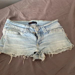 Ljusa jeansshorts från Crocker - Säljer ett par ljusa jeansshorts från Crocker i modellen CR Superslim TB 50. De har en snygg, sliten look med fransiga kanter och en låg midja. Perfekta för sommaren och ger en avslappnad stil. Storlek 27, vilket motsvarar dam XS.