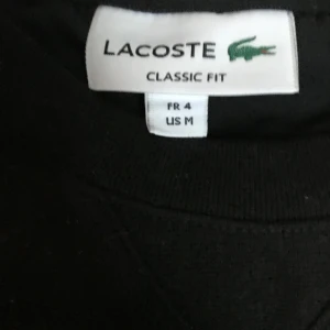 Svart tröja från Lacoste - Säljer en klassisk svart tröja från Lacoste i storlek M. Tröjan har en enkel design med det ikoniska Lacoste-logotypen på bröstet. Den är långärmad och har en normal passform, perfekt för både vardag och avslappnade tillfällen.
