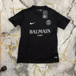Svart PSG x Balmain fotbollströja - Säljer en snygg svart PSG x Balmain fotbollströja från Nike. Tröjan har en stilren design med PSG-loggan och Balmain Paris tryckt på framsidan. Den är kortärmad och tillverkad i ett bekvämt Dri-FIT material som håller dig sval. Perfekt för både träning och vardag!