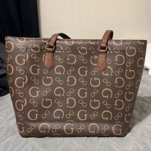 Guess väska - Väskan är helt ny och är äkta. Den har inga som helst skador. Det finns en zip ficka inuti samt ett litet fack. Det finns ett till fack utanpå väskan som man också kan använda. Den är köpt i en Guess butik i Usa.