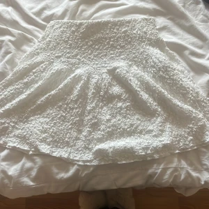 Vit kjol med struktur - Säljer en superfin vit kjol med en härlig struktur i tyget. Den har en elastisk midja som gör den bekväm att bära. Perfekt för sommardagar eller en festlig kväll. Kjolen är i nyskick och redo för nya äventyr!