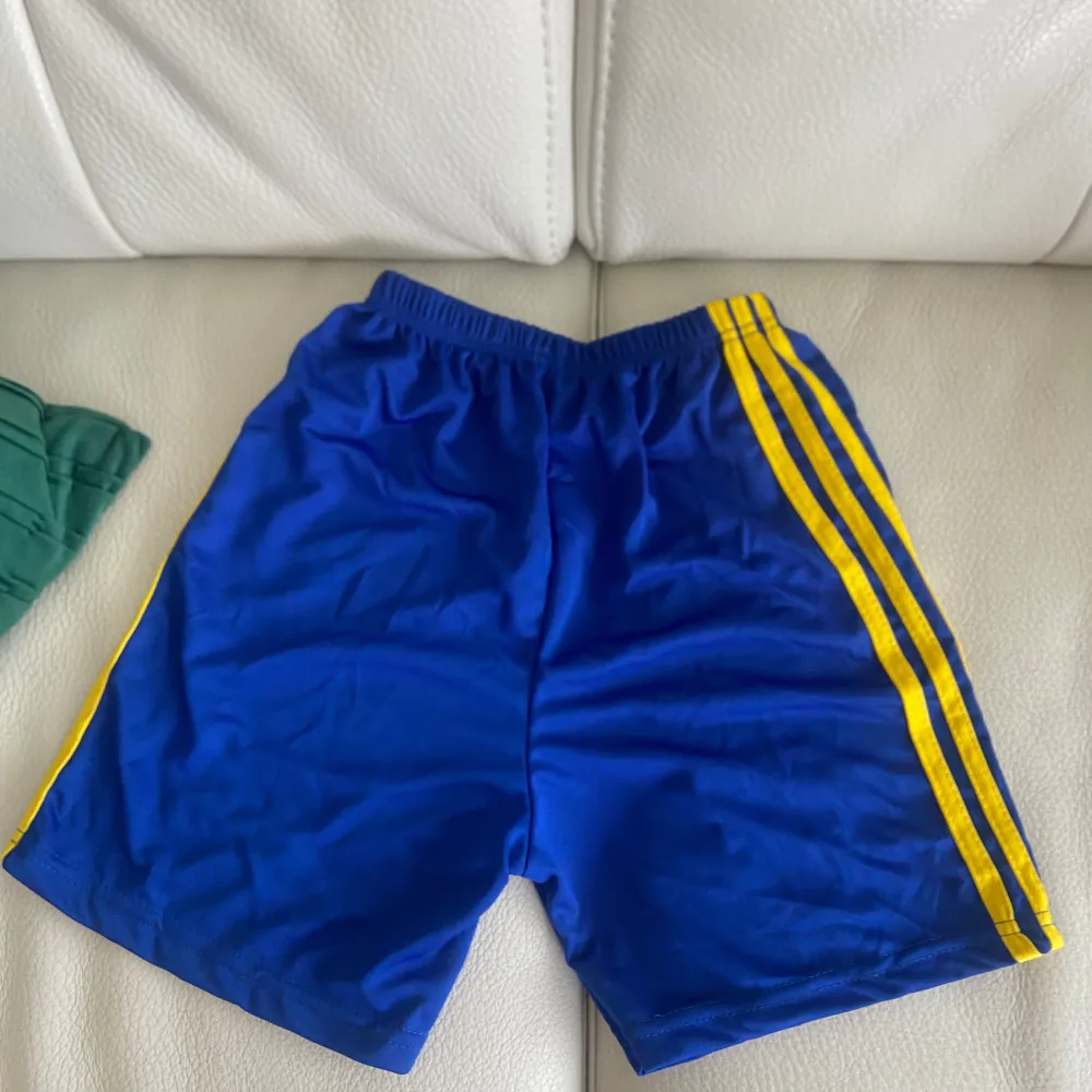 Säljer ett par blå fotbollsshorts från Adidas med gula ränder längs sidorna. Shortsen har en elastisk midja och ett tryckt emblem på ena benet. Perfekta för fotbollsträningen eller matchen! Har aldrig används säljer för att har två likadana (Lamine yamal). Shorts.