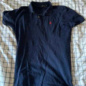 Mörkblå pikétröja från Ralph Lauren - Säljer en klassisk mörkblå pikétröja från Ralph Lauren. Den har en röd broderad logga på bröstet och är i storlek M. Perfekt för en stilren och avslappnad look. Tröjan är i bra skick och redo för nya äventyr!