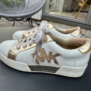 Vita och guldiga sneakers från Michael Kors - Säljer ett par snygga sneakers från Michael Kors i vitt och guld. Skorna har en stilren design med guldiga detaljer och snörning framtill. De är perfekta för både vardag och festliga tillfällen. Michael Kors-loggan syns tydligt på sidan och bak på skorna.