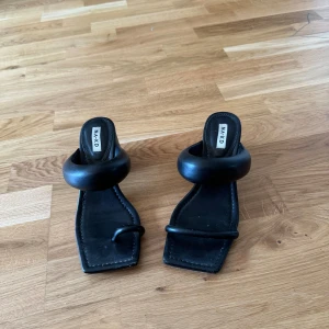 Svarta klackskor från NA-KD - Säljer ett par stilrena svarta klackskor från NA-KD. De har en fyrkantig tå och en bred rem över foten. Klacken är stabil och ger en elegant look. Perfekta för både vardag och fest!