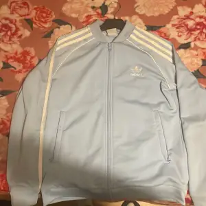 Säljer en snygg ljusblå Adidas jacka med de klassiska tre ränderna på ärmarna. Jackan har dragkedja framtill och två fickor. Perfekt för en sportig look eller bara för att chilla. Adidas-loggan är broderad på bröstet. Passar perfekt för vår och höst!