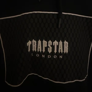 Svart hoodie från Trapstar London - Säljer en svart hoodie från Trapstar London. Den har en cool design med ett mönstrat tryck på framsidan och texten 'Trapstar London'. Perfekt för en avslappnad och stilren look. Hoodien är använd 1 gång. Nypris är 150£.