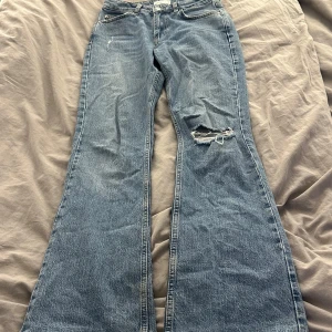 Blå jeans med slitningar - Säljer ett par snygga blå jeans från Perfect Jeans i storlek S. De har en cool slitning på ena benet och en klassisk femficksdesign. Jeansen har en bootcut-stil som ger en retro vibe. Perfekta för en avslappnad look! ”Har trampat lite på de”