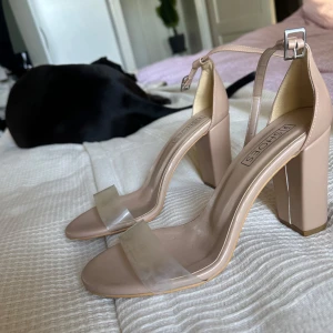 Beige klackskor med transparent rem - Säljer ett par snygga beige klackskor från Shoes. De har en stilren design med en bred transparent rem över tårna och en justerbar rem runt ankeln. Perfekta för sommarens fester eller en elegant kväll ute. Klacken är stabil och ger bra stöd.