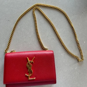 Röd axelväska från YSL - Säljer en superfin röd axelväska från YSL. Väskan är i skinn och har en elegant guldfärgad kedja som axelrem. Den har en snygg YSL-logga i guld på framsidan. Köpt i Stockholm, knappt använd.