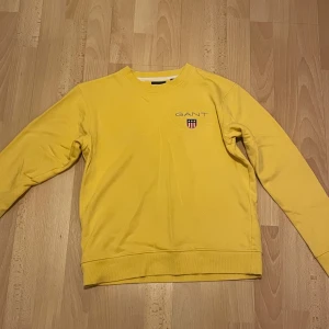 Gul tröja från GANT - Säljer en snygg gul tröja från GANT. Den har en klassisk design med rund hals och långa ärmar. Perfekt för en avslappnad stil och passar bra till både jeans och chinos. GANT-loggan är broderad på bröstet för en stilren look.