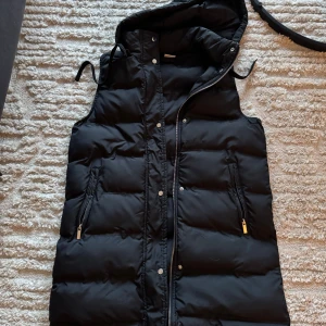 Svart dunväst med huva - Säljer en snygg och praktisk svart dunväst med huva. Västen har en dragkedja och knappar framtill samt två fickor med dragkedja. 