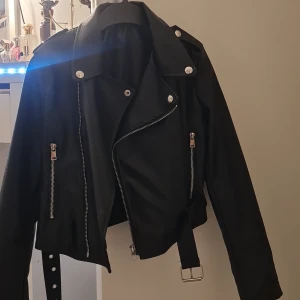 Svart skinnjacka med dragkedjor - Säljer en snygg svart skinnjacka.. Den är endast använd 1 gång till en tema grej. Det är ett bälte i midjan för extra edge. Och dragkedja längst ner på armarna. Perfekt för höst och vår, och funkar lika bra till fest som till vardags. Pm för mer info