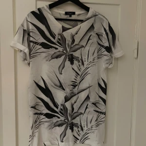 Vit och svart t-shirt med bladmönster från New Look - Säljer en snygg vit t-shirt från New Look med ett coolt svart bladmönster. Den har korta ärmar och en normal passform, perfekt för en avslappnad stil. Passar bra till både jeans och shorts!