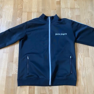 Svart jacka från Palm Angels - Säljer en svart jacka från Palm Angels med dragkedja framtill och vita detaljer. Jackan har långa ärmar och en stilren design med logotypen tryckt på bröstet. Perfekt för en casual look!