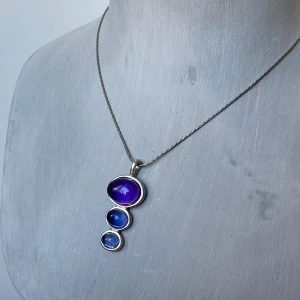 Halsband med blå och lila stenar - Säljer ett snyggt halsband med tre ovala stenar i olika storlekar. Stenarna är i nyanser av blått och lila, infattade i en silverram. Kedjan är tunn och elegant, perfekt för både vardag och fest. Låset är justerbart för att passa olika längder. Okänt material 