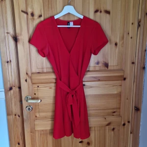 Röd omlottklänning - Säljer en superfin röd omlottklänning med korta ärmar. Den har en v-ringad hals och ett knytband i midjan som ger en snygg passform. Perfekt för sommarens alla fester eller varför inte på juldagen!