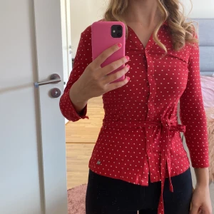Röd prickig blus med knytband - Säljer en superfin röd blus med vita prickar. Den har långa/trekvart ärmar och knappar framtill. Blusen kommer med ett knytband i midjan för en snygg passform. Perfekt för både vardag och fest!