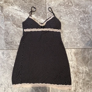Svart prickig klänning med spetsdetaljer - Säljer ett sött svart nattlinne med vita prickar och fina spetsdetaljer. Den har smala axelband och en v-ringad design med spets längs kanterna. 