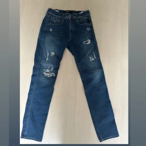 Blå jeans från Replay - Säljer ett par snygga blå jeans från Replay, modell Anbass. De har en cool slitningseffekt och är i en straight passform. Perfekta för en avslappnad stil!