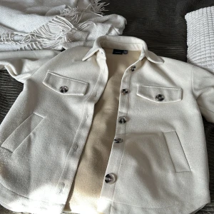 Vit overshirt med knappar - Säljer en stilren vit overshirt med stora knappar. Den har två bröstfickor och två sidofickor, perfekt för en casual look. Materialet känns mjukt och bekvämt, perfekt för lager-på-lager under hösten.