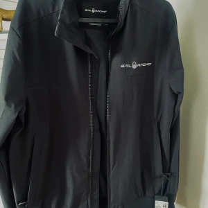 Svart jacka från Sail Racing - Säljer en snygg svart jacka från Sail Racing. Den har en stilren design med dragkedja och logotyp på bröstet. Jackan är perfekt för både vår och höst, och har långa ärmar. Materialet känns slitstarkt och av hög kvalitet. Passar perfekt för en casual look eller när du vill ha något bekvämt men ändå stiligt.  Jackan är oanvänd. 