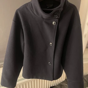 Svart kappa från Bershka - Säljer en stilren svart kappa från ginatricot i storlek XS. Den har en hög krage och knäpps med knappar framtill. Perfekt för höst och vår, med en klassisk look som funkar till allt. Jackan är i mycket bra skick och redo för nya äventyr!