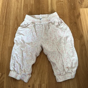 Vita byxor med blommönster från Pippi - Söta vita byxor från Pippi med ett diskret blommönster i rosa och grönt. Byxorna har elastisk midja och muddar vid bensluten för extra komfort. Perfekta för små barn som vill ha något bekvämt och stiligt.