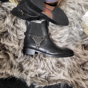 Svarta boots från Tommy Hilfiger - Snygga svarta boots i skinn från Tommy Hilfiger med dekorativt bälte och metallspänne. Perfekta för en stilren look. De har bara legat i garderoben och är i storlek 40...Jag säljer dem för 1000 kr