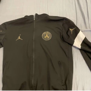 Svart träningsjacka från Jordan x PSG - Säljer en snygg svart träningsjacka från Jordan i samarbete med PSG. Jackan har en dragkedja framtill och är långärmad med vita detaljer på ärmarna. Perfekt för fotbollsträning eller som en stilren vardagsjacka. Den är i mycket bra skick och redo för nya äventyr!