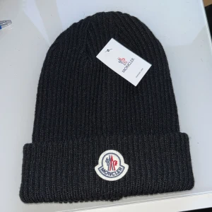 Svart ribbad mössa från Moncler - Säljer en snygg svart ribbad mössa från Moncler i nytt skick. Den är perfekt för vintern och har en stilren design med Moncler-loggan framtill. Mössan är i one size och passar de flesta. Håll dig varm och trendig i kylan!