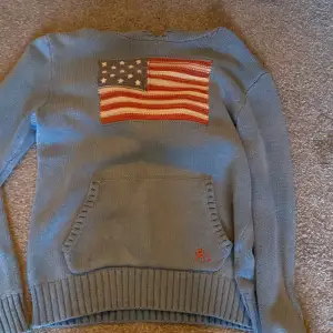 Säljer en snygg blå stickad tröja från Polo Ralph Lauren med en stor amerikansk flagga på framsidan. Tröjan har en praktisk ficka framtill och RL-broderi i rött. Perfekt för kyliga dagar och ger en klassisk look. Passar både till vardags och lite finare tillfällen.