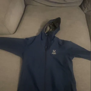 Blå vindjacka från Haglöfs - Säljer en blå vindjacka från Haglöfs i storlek S. Jackan har en huva och dragkedja framtill. Perfekt för blåsiga dagar och utomhusaktiviteter. Den är i bra skick och redo för nya äventyr!