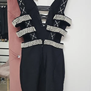 Svart klänning med pärldetaljer  - Snygg svart klänning med djup v-ringning och unika pärldetaljer. Klänningen har en tight passform och är dekorerad med pärlor i ett geometriskt mönster. Perfekt för en kväll ute!