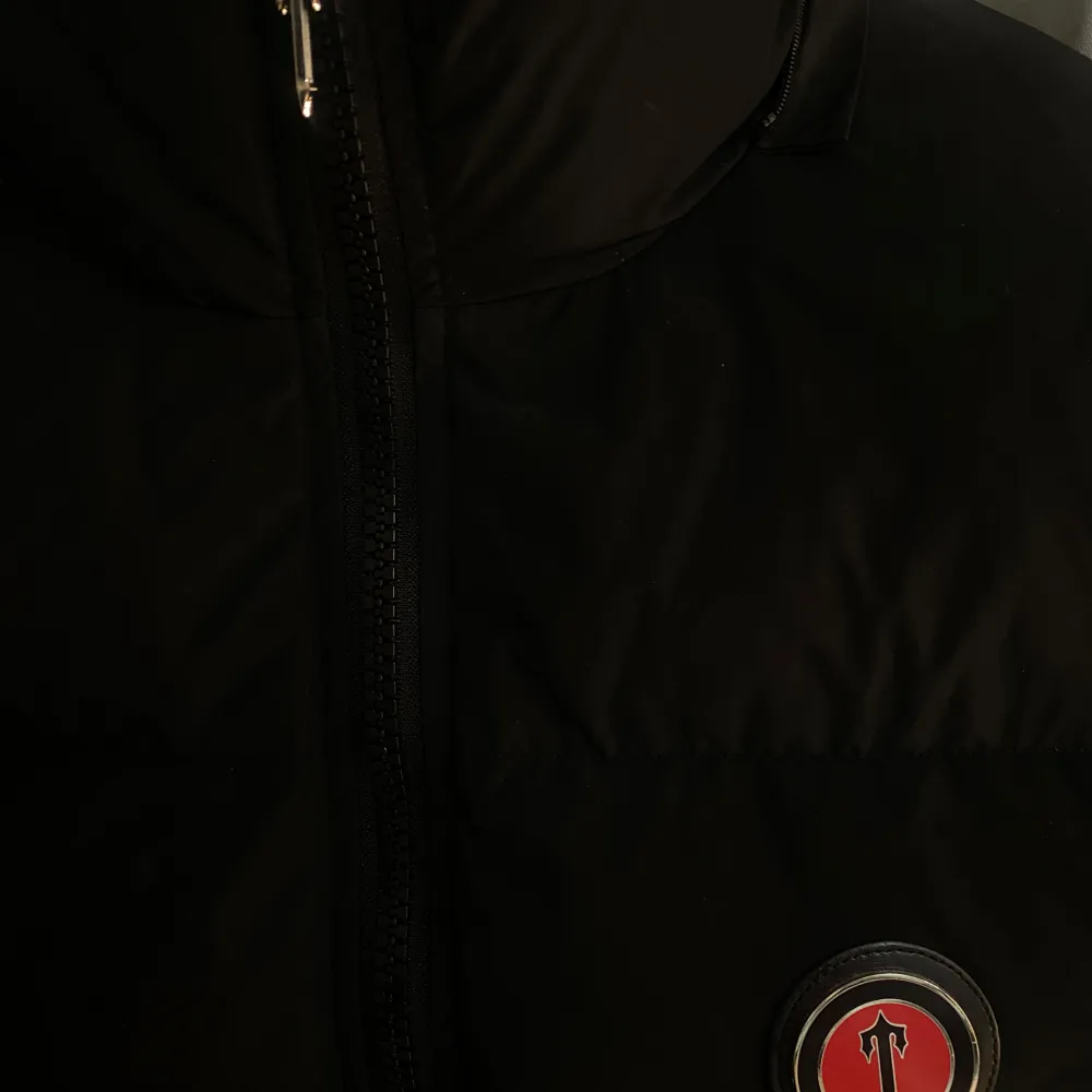 Snygg svart pufferjacka från Trapstar med dragkedja och broderad logga på bröstet. Jackan har en hög krage och texten 'Trapstar' i rött på ryggen. Perfekt för kyliga dagar.. Takit.