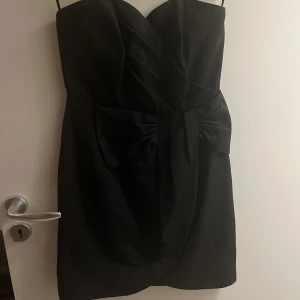 Svart axelbandslös klänning - Elegant svart axelbandslös klänning med en stor rosett framtill. Perfekt för festliga tillfällen. Klänningen har en figurnära passform och är kort i modellen.