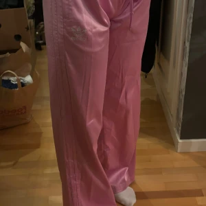 Rosa byxor från Adidas - Snygga rosa träningsbyxor från Adidas med klassiska tre ränder längs sidorna. Byxorna har en loose passform och är dekorerade med Adidas-loggan i strass på benet. Det är mycket sparsamt använda 