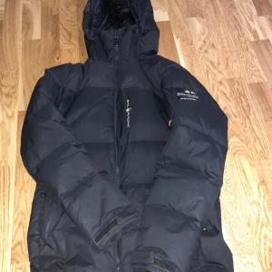 Svart dunjacka från Sail Racing - Säljer en svart dunjacka från Sail Racing i mycket bra skick. Jackan har en cool expedition-stil med broderad logga och dragkedja framtill. Perfekt för kalla vinterdagar med sin tjocka vaddering och justerbara huva. Den är både snygg och funktionell, idealisk för vinteräventyr! ❄️