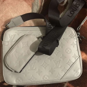 Grå axelväska från Louis Vuitton - Snygg grå axelväska från Louis Vuitton med det klassiska monogrammönstret. Väskan har en justerbar axelrem och en praktisk dragkedja. Perfekt för att bära dina nödvändigheter med stil.